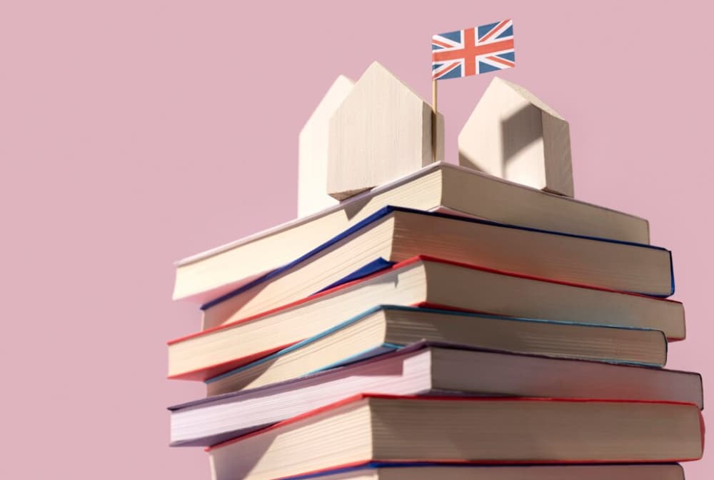 Books stacked with a toy house and UK flag on top, pink backdrop